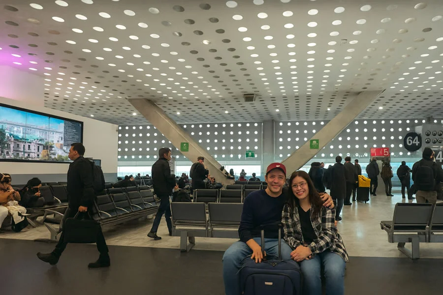 Pareja sonriente sentada en la zona de embarque de la Terminal 2 del Aeropuerto de la Ciudad de México con su equipaje, esperando la salida de su vuelo